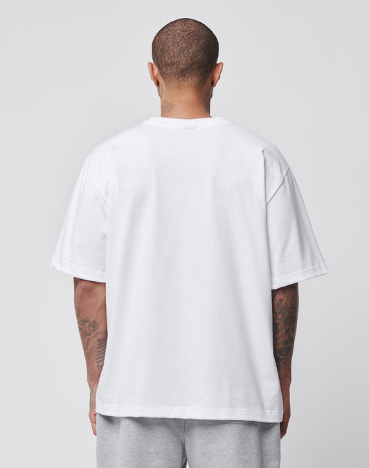 A person with short hair and visible arm tattoos stands facing away from the camera, wearing a plain white LIVE FAST DIE YOUNG LFDY x Trigema Tennis Tee and light gray shorts against a simple light background.