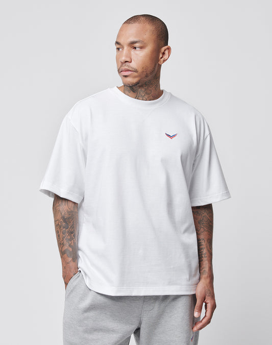 A tattooed man wears a LIVE FAST DIE YOUNG LFDY x Trigema Tennis Tee in white, Oversize Fit with a small left chest emblem, paired with light gray sweatpants, standing against a plain light gray background and looking to his left.