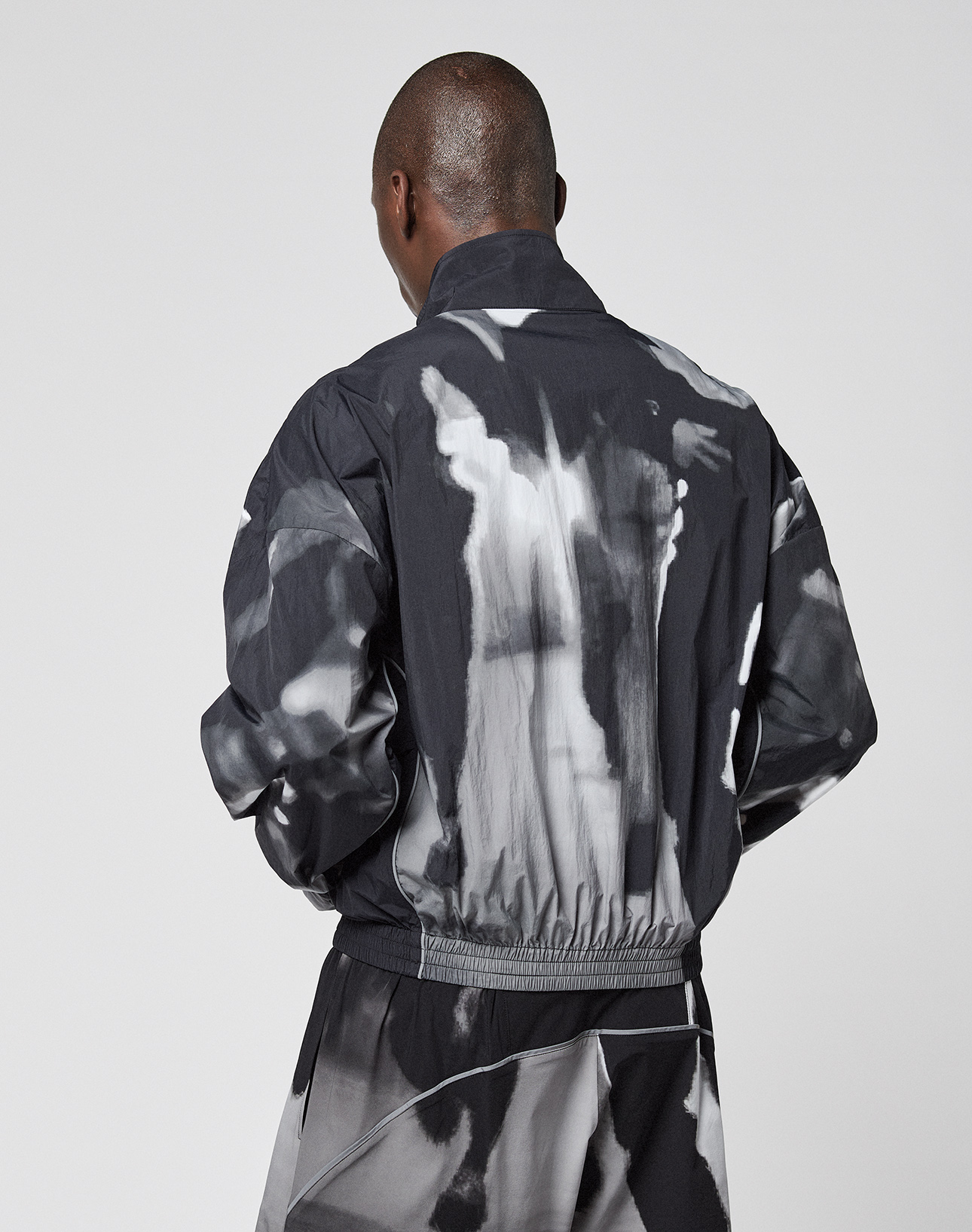 A person with short hair stands with their back to the camera, wearing the black and white LFDY Performance Jacket by LIVE FAST DIE YOUNG, featuring an allover abstract print, against a plain light background.