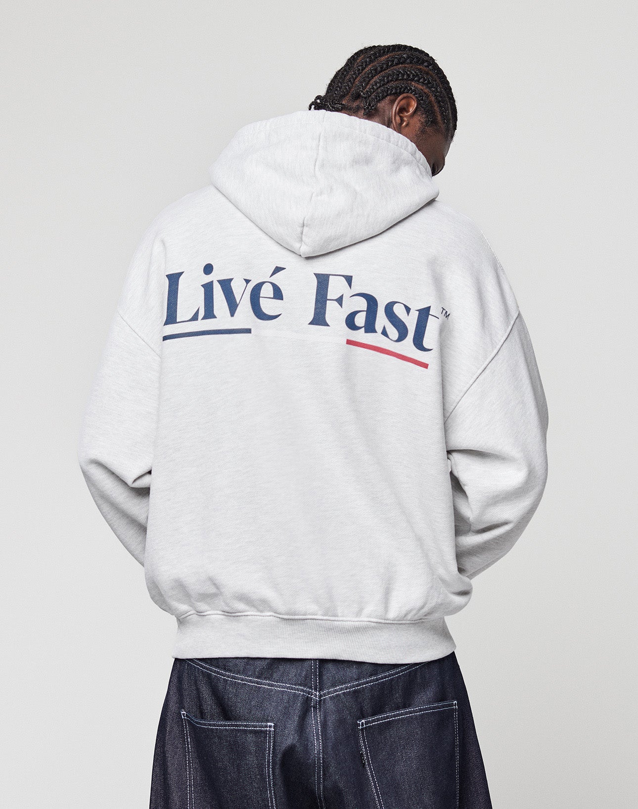 A person with braided hair, facing away, wears an oversized LIVE FAST DIE YOUNG Live Fast Paris Hooded hoodie with a bold blue and red backprint, paired with dark jeans against a plain light background.