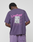 A person faces away, wearing the LIVE FAST DIE YOUNG Basic Evidence Tee in Oversize Fit purple with "LiveFast" printed in pink and white on the back and a silver rectangle patch. Made from 100% Baumwolle.