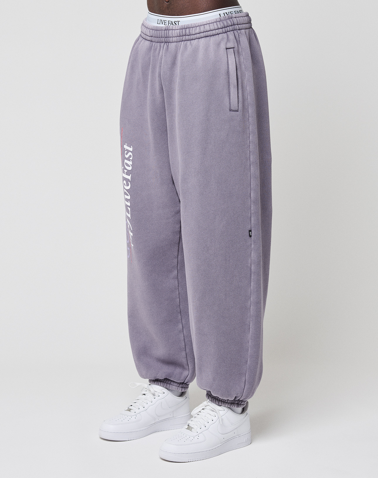 A person wears LIVE FAST DIE YOUNG’s LFDY Hotline Loose Jogger in washed lavender with an elastic waistband, leg print reading PAST and LOST, and elastic cuffs. Styled with white sneakers against a plain white background.