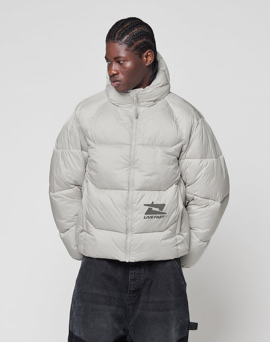 A person with braided hair wears the LIVE FAST DIE YOUNG Live Fast Puffer Jacket in light gray with a high collar and reflective "LIVE FAST" print, paired with dark pants, standing against a plain white background.