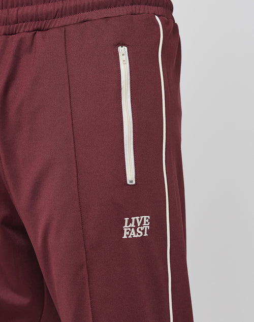 Close-up of maroon Live Fast Trackjogger by LIVE FAST DIE YOUNG, featuring a Regular Fit, white zippered pocket, white side piping, and “LIVE FAST” logo embroidered in white below the pocket.