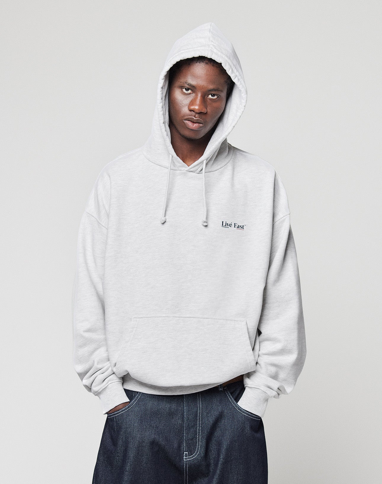 Someone stands against a plain light background, hands in the front pocket, wearing an oversized fit, light gray LIVE FAST DIE YOUNG Live Fast Paris Hooded with front and back print, looking directly at the camera.