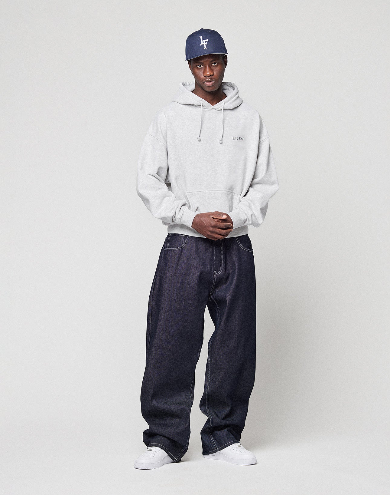 A man stands against a plain background, wearing a navy baseball cap, the LIVE FAST DIE YOUNG Live Fast Paris Hooded pullover in light gray, dark denim jeans, and white sneakers. His hands are clasped as he looks at the camera.