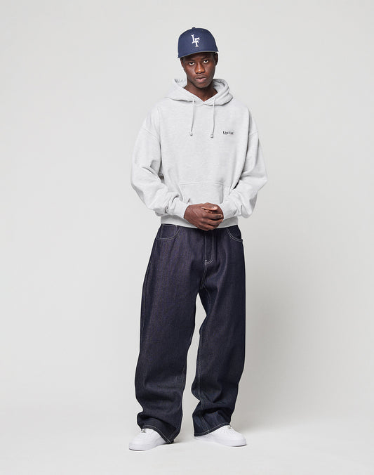 A man stands against a plain background, wearing a navy baseball cap, the LIVE FAST DIE YOUNG Live Fast Paris Hooded pullover in light gray, dark denim jeans, and white sneakers. His hands are clasped as he looks at the camera.