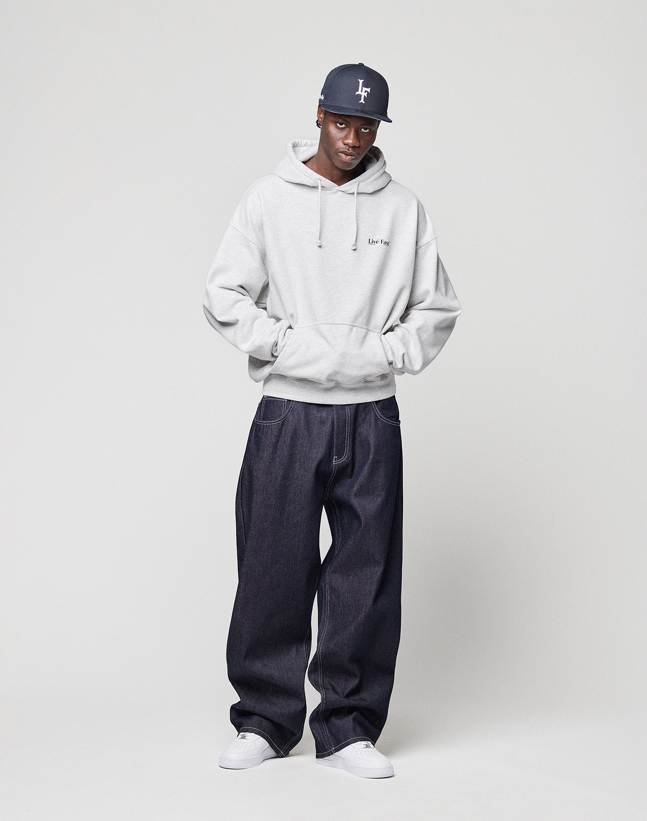 A person wears an oversized Live Fast Paris Hooded by LIVE FAST DIE YOUNG with front and back print, paired with dark baggy jeans, white sneakers, and a gray cap, standing against a plain background with hands in pockets.