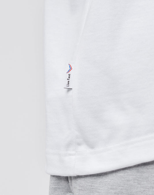 Close-up of the LFDY x Trigema Tennis Tee by LIVE FAST DIE YOUNG, a white oversize-fit t-shirt with a small side tag featuring red and blue accents and Live Fast text, styled over light gray pants against a plain background.