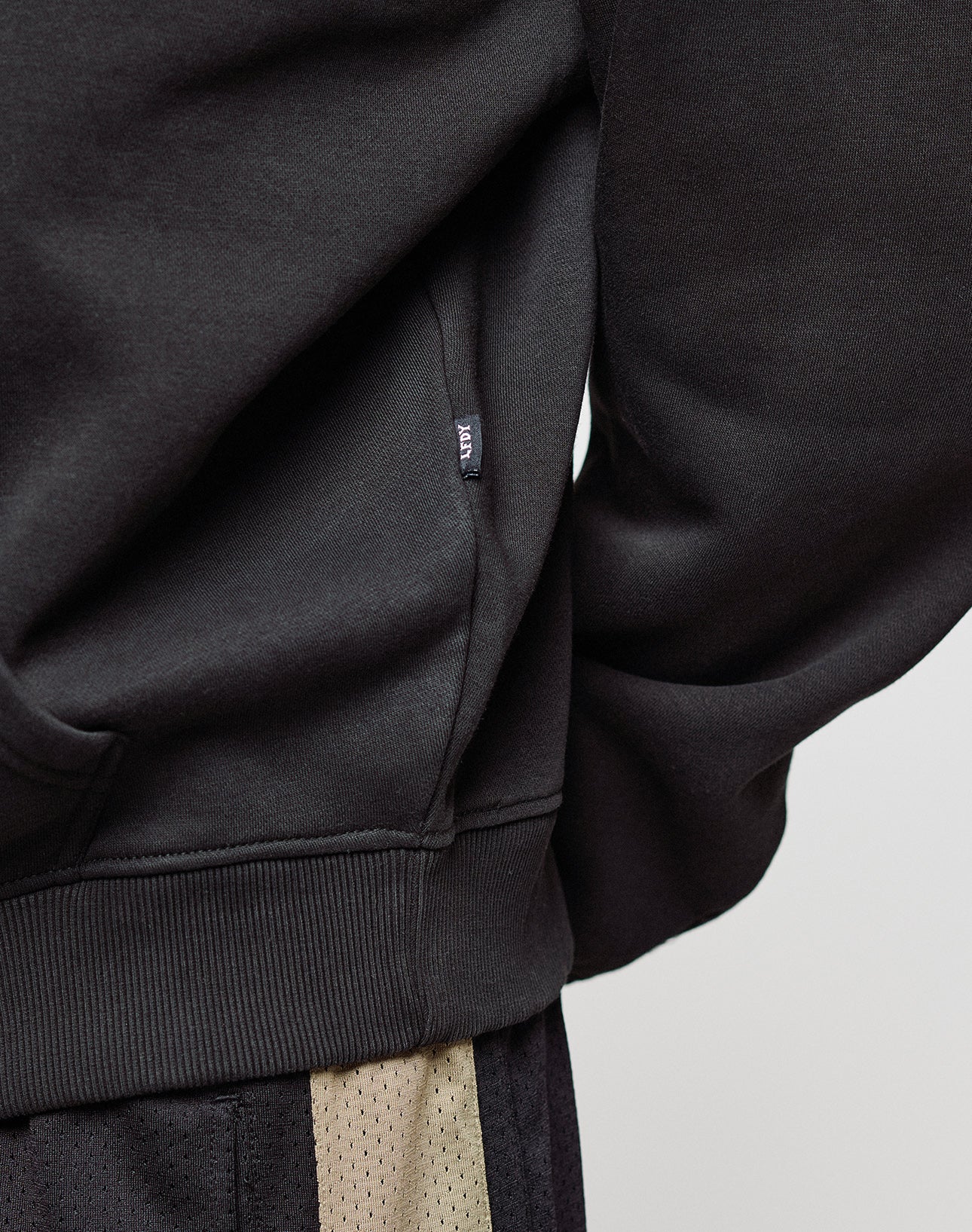 Close-up of a person wearing the LIVE FAST DIE YOUNG Basic Blank Hooded sweatshirt in black and black and beige mesh athletic shorts. A small black Flag-Label appears on the side seam. The background is plain and light-colored.