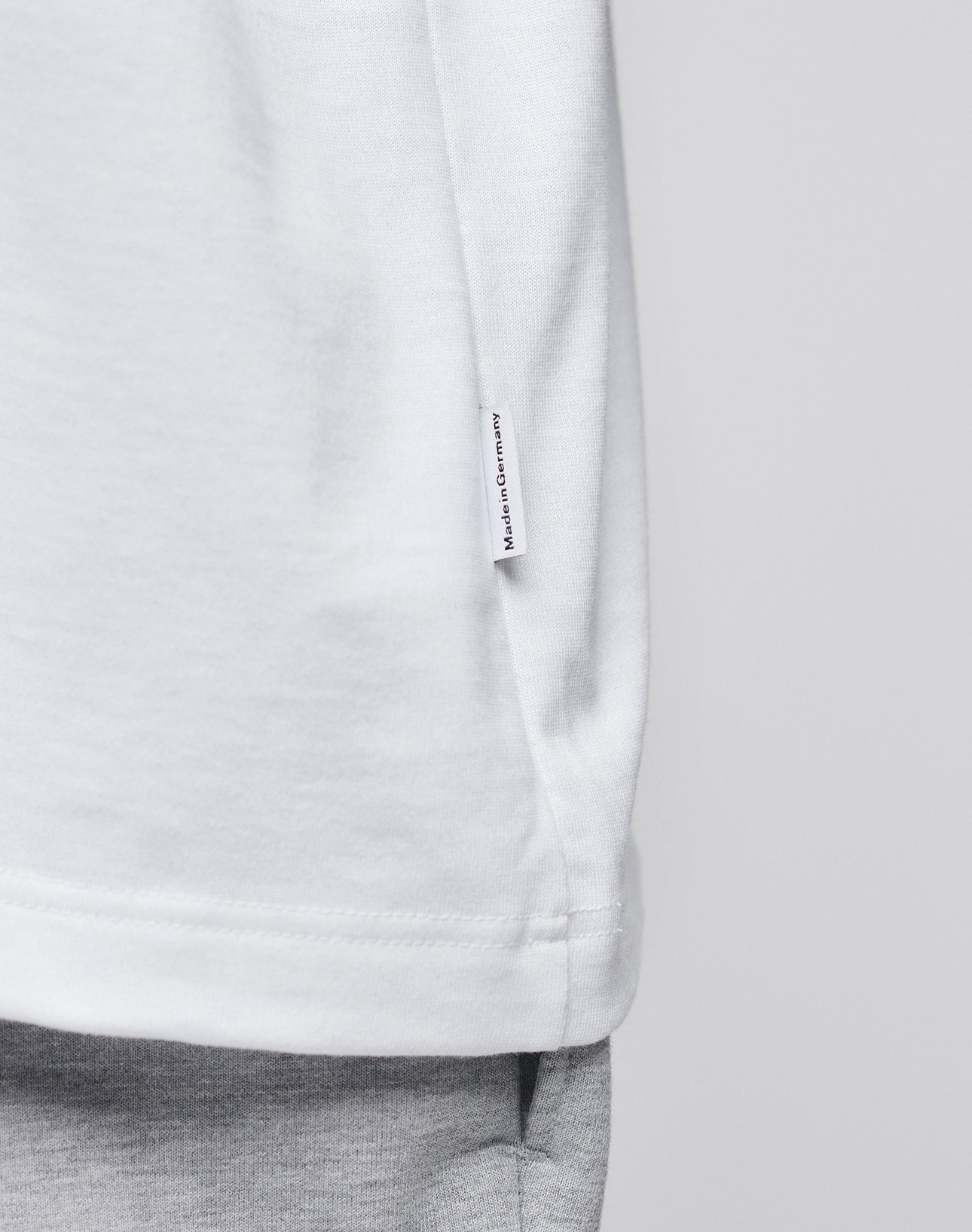 Close-up of the LFDY x Trigema Tennis Tee by LIVE FAST DIE YOUNG—an oversize white T-shirt with a “Hergestellt in Deutschland” tag on the side seam, styled with light gray pants against a plain, neutral background.