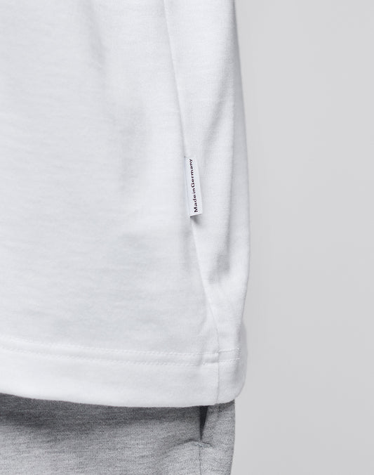 Close-up of the LFDY x Trigema Tennis Tee by LIVE FAST DIE YOUNG—an oversize white T-shirt with a “Hergestellt in Deutschland” tag on the side seam, styled with light gray pants against a plain, neutral background.