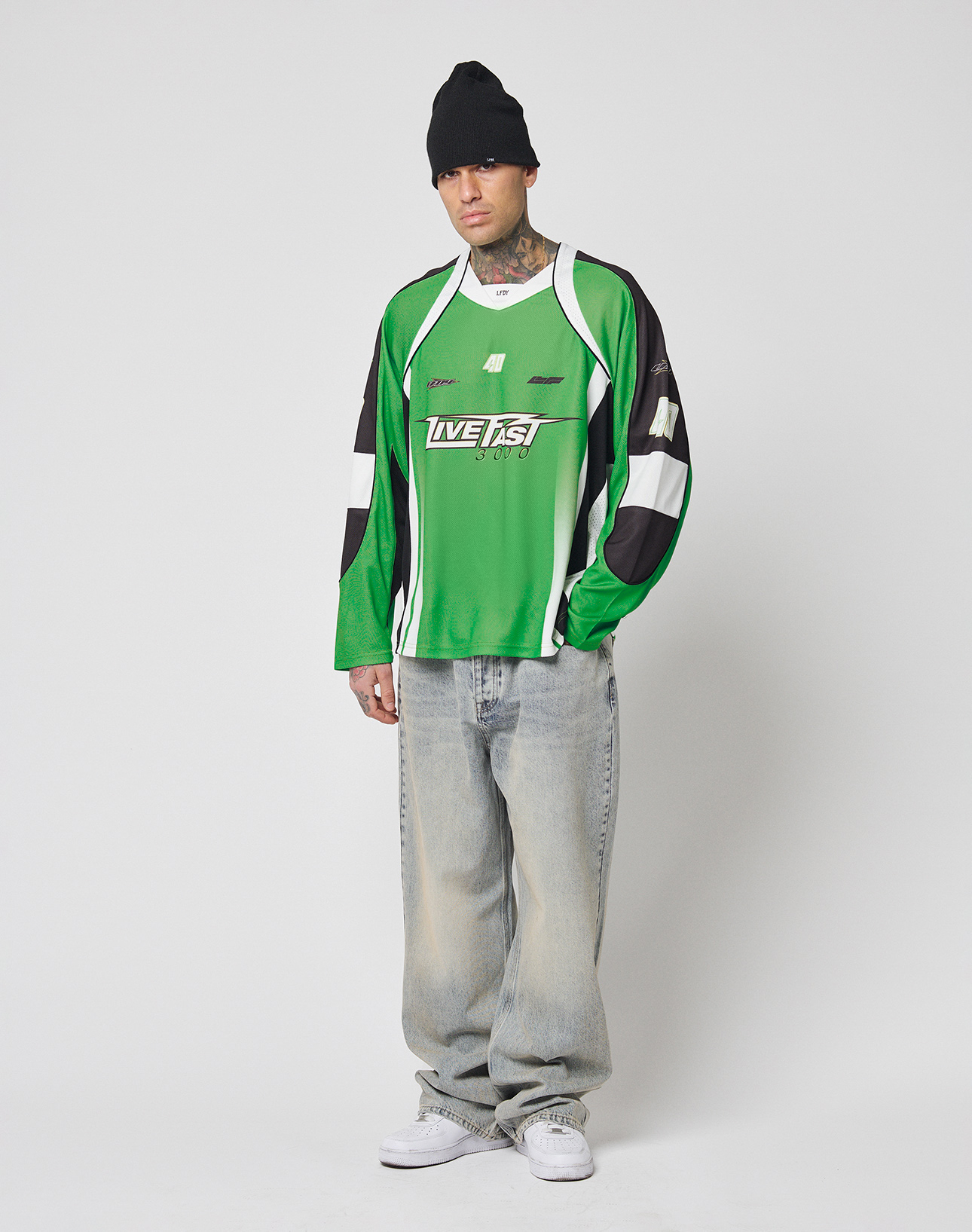 A person in a black beanie, baggy light-wash jeans, white sneakers, and the LIVE FAST DIE YOUNG LFDY Motorcross Jersey with green, black, and multi-logo prints stands against a plain white background.