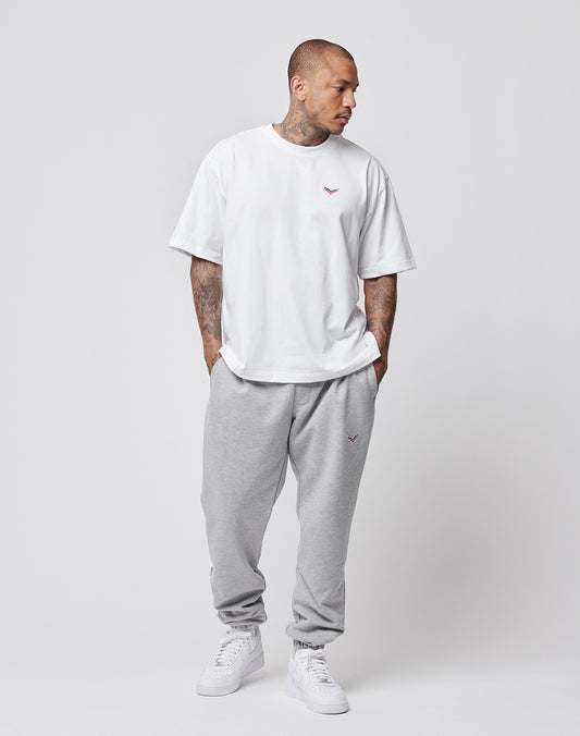 A person wearing the LIVE FAST DIE YOUNG LFDY x Trigema Tennis Tee, loose light gray sweatpants, and white sneakers stands against a plain light background, looking left with hands in pockets.