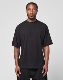 A man with braided hair and a short beard faces forward, wearing a loose black LFDY Vintage Tee by LIVE FAST DIE YOUNG and black pants. He shows visible tattoos on his right arm and poses against a plain light background.