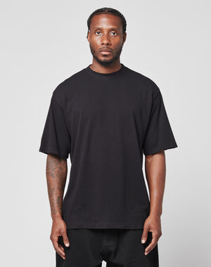 A man with braided hair and a short beard faces forward, wearing a loose black LFDY Vintage Tee by LIVE FAST DIE YOUNG and black pants. He shows visible tattoos on his right arm and poses against a plain light background.