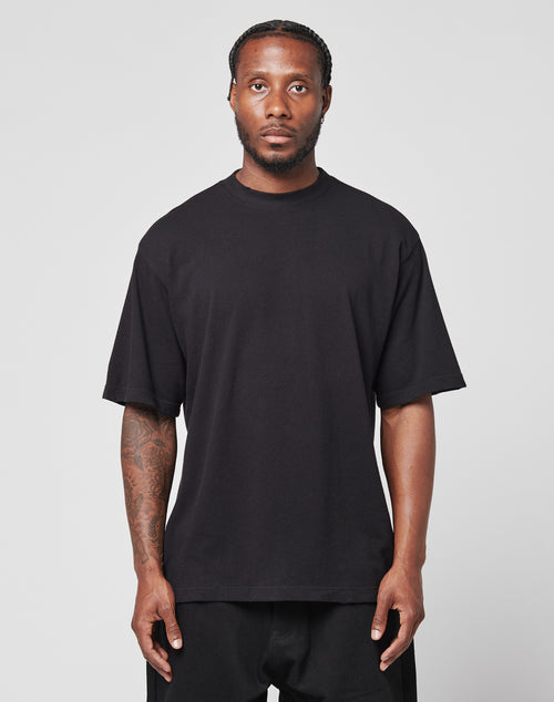 A man with braided hair and a short beard faces forward, wearing a loose black LFDY Vintage Tee by LIVE FAST DIE YOUNG and black pants. He shows visible tattoos on his right arm and poses against a plain light background.