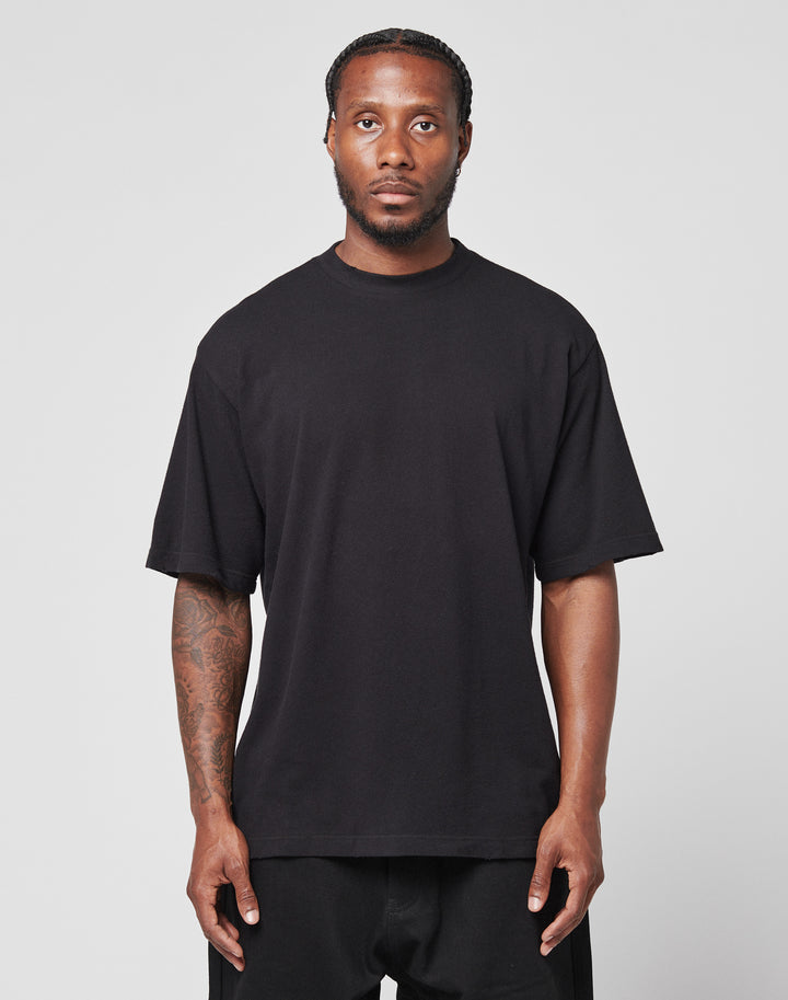 A man with braided hair and a short beard faces forward, wearing a loose black LFDY Vintage Tee by LIVE FAST DIE YOUNG and black pants. He shows visible tattoos on his right arm and poses against a plain light background.