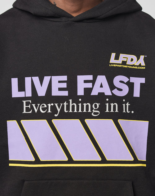 A person wears the LFDY Racing Hooded by LIVE FAST DIE YOUNG: an oversized black hoodie with glossy LFDA LIVE FAST Everything in it. print in purple and white, a yellow-striped graphic, and LIVEFASTDIEYOUNG.COM on the chest.