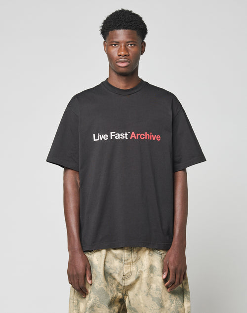 A young man stands against a plain background, wearing the LIVE FAST DIE YOUNG Live Fast Archive Tee—an oversize fit black t-shirt with bold white and red front print—paired with light tan camouflage pants. Crafted from premium Baumwolle.