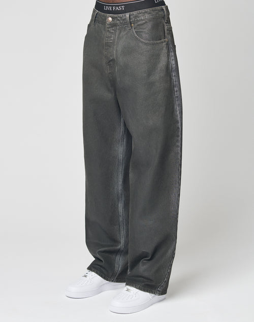 A person in LIVE FAST DIE YOUNG’s LFDY Coated Baggy Denim—baggy, wide-leg, dark grey 100% Baumwolle jeans—paired with white sneakers and LIVE FAST waistband black underwear stands against a plain white background.
