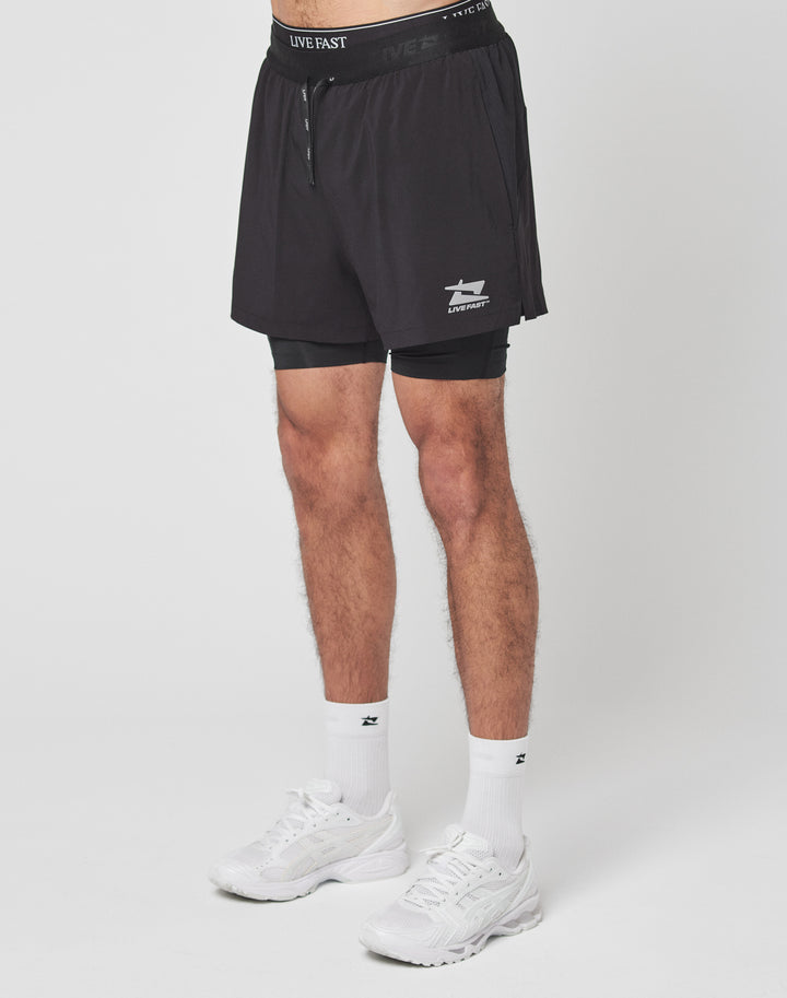A person models LIVE FAST DIE YOUNG Live Fast Performance Running Shorts in black with integrierte Tights and a small white logo, paired with white crew socks and athletic shoes. The shot only shows the shorts; the persons torso and head are not visible.
