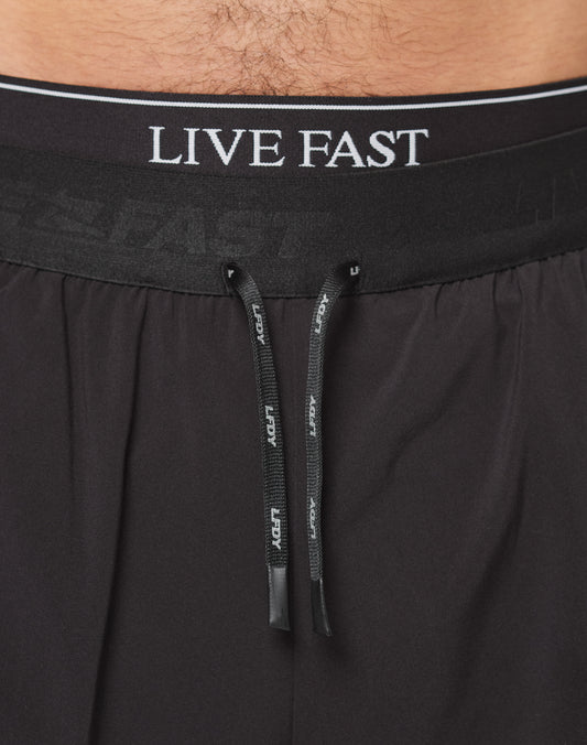 Close-up of a person’s lower torso wearing Live Fast Performance Running Shorts by LIVE FAST DIE YOUNG. The black shorts have an elastic waistband with white LIVE FAST branding and black drawstrings with white text in front.