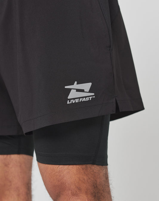A close-up shows a person wearing black Live Fast Performance Running Shorts by LIVE FAST DIE YOUNG, featuring integrated tights and a prominent LIVE FAST logo on the left leg with part of their left and right thighs visible.