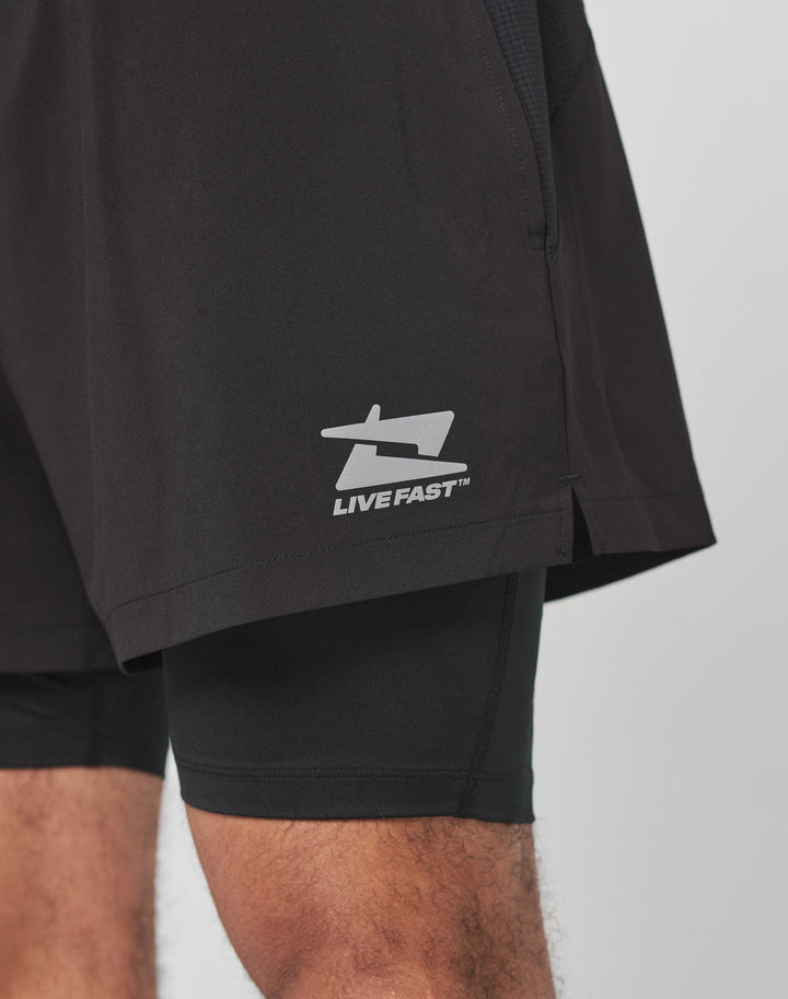 A close-up shows a person wearing black Live Fast Performance Running Shorts by LIVE FAST DIE YOUNG, featuring integrated tights and a prominent LIVE FAST logo on the left leg with part of their left and right thighs visible.