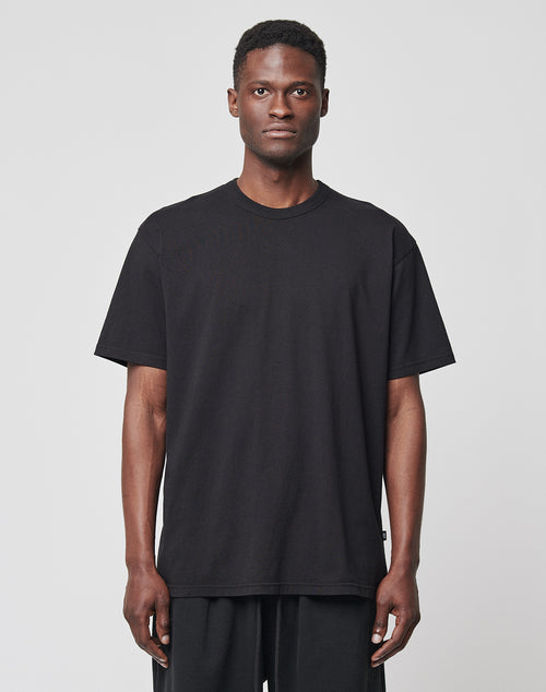 A man with short hair and a neutral expression stands facing forward against a plain background, wearing the LIVE FAST DIE YOUNG LFDY Blank Tee in black and matching black pants. The T-shirt is regular fit and made from cotton.