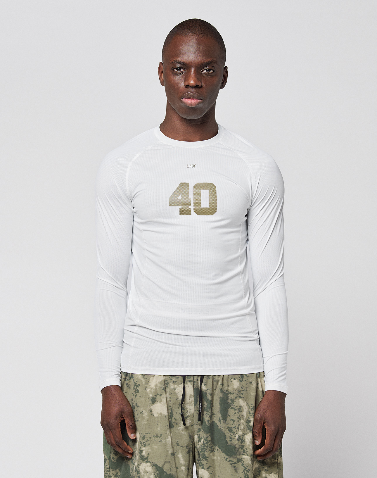 A person faces forward in a LIVE FAST DIE YOUNG LFDY Performance Slim Longsleeve, white with the number 40 on the front, and green-gray camouflage shorts made from leichtes Obermaterial, set against a plain light background.