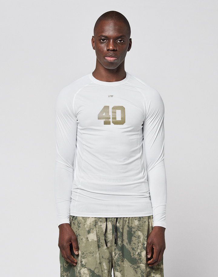 A person faces forward in a LIVE FAST DIE YOUNG LFDY Performance Slim Longsleeve, white with the number 40 on the front, and green-gray camouflage shorts made from leichtes Obermaterial, set against a plain light background. | model