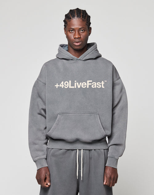 A man faces the camera in a gray LIVE FAST DIE YOUNG Basic +49 Hooded sweatshirt and matching sweatpants, featuring a bold white "+49LiveFast" front print. The background is plain and light-colored.