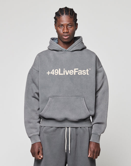 A man faces the camera in a gray LIVE FAST DIE YOUNG Basic +49 Hooded sweatshirt and matching sweatpants, featuring a bold white "+49LiveFast" front print. The background is plain and light-colored.