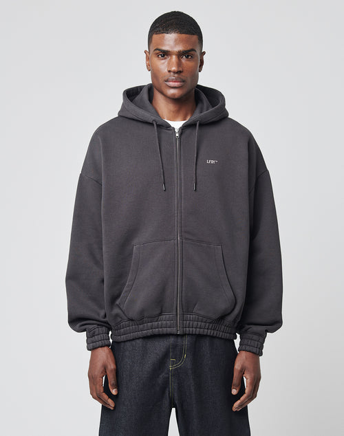 A man stands against a plain light gray background, wearing the LIVE FAST DIE YOUNG Die Young Hooded Zip—a black oversize fit hoodie with front and back print, hood, front pockets, hidden zip, and a small white logo on the left chest—paired with dark jeans.