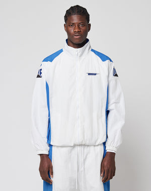 A man faces forward against a plain light gray background, wearing the LIVE FAST DIE YOUNG LFDY Trackjacket 3000—white and blue with a high collar, zip-up front, and matching pants featuring Multilogo Stick details.