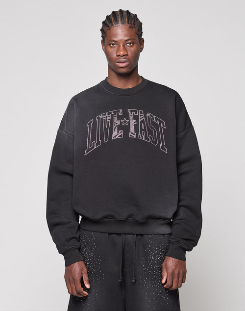 Against a plain background, a person wears the LIVE FAST DIE YOUNG Live Fast Rhinestone Sweater in an oversized fit with "LIVE FAST" on the front, paired with matching black pants featuring subtle white speckles.