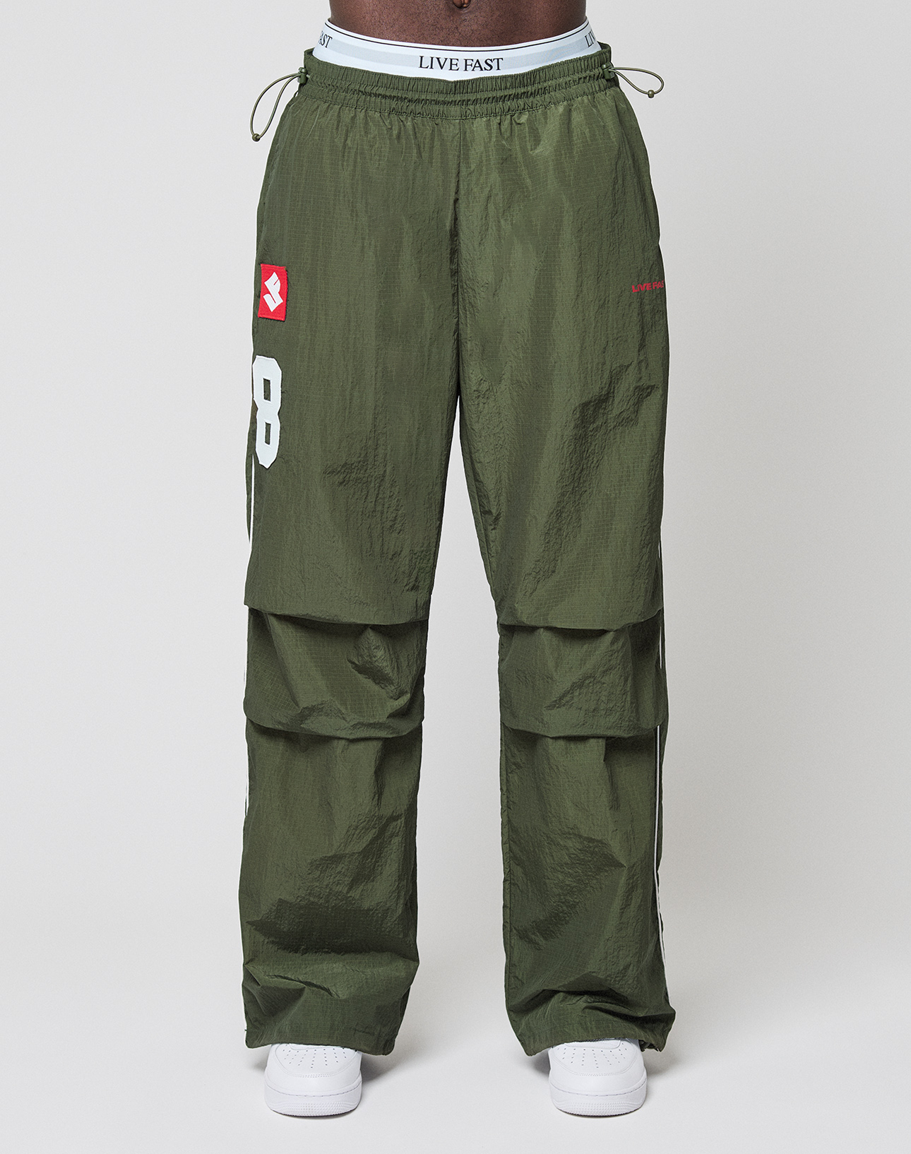 A person wears LFDY Nylon Trackpants by LIVE FAST DIE YOUNG, featuring olive green wide fit, white logo-print numbers with red accents, and a LIVE FAST waistband, paired with white sneakers against a plain white background.