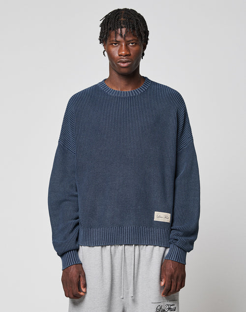 A man with short, twisted hair wears the LIVE FAST DIE YOUNG LFDY Knit Sweater in blue and light gray sweatpants, standing against a plain light gray background.