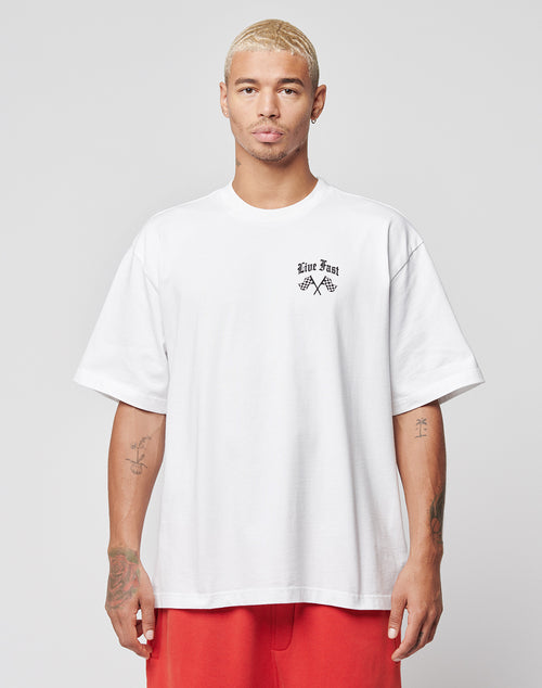 A person with short blond hair wears the LFDY Haters Tee by LIVE FAST DIE YOUNG in oversize fit, featuring Live Fast text and chest graphics, paired with red pants and standing against a plain light gray background.