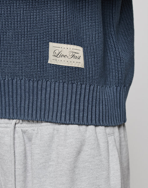 A close-up of the LFDY Knit Sweater by LIVE FAST DIE YOUNG in blue 100% Baumwolle, featuring a beige "Live Fast" patch and dropped shoulders, styled with light grey sweatpants.
