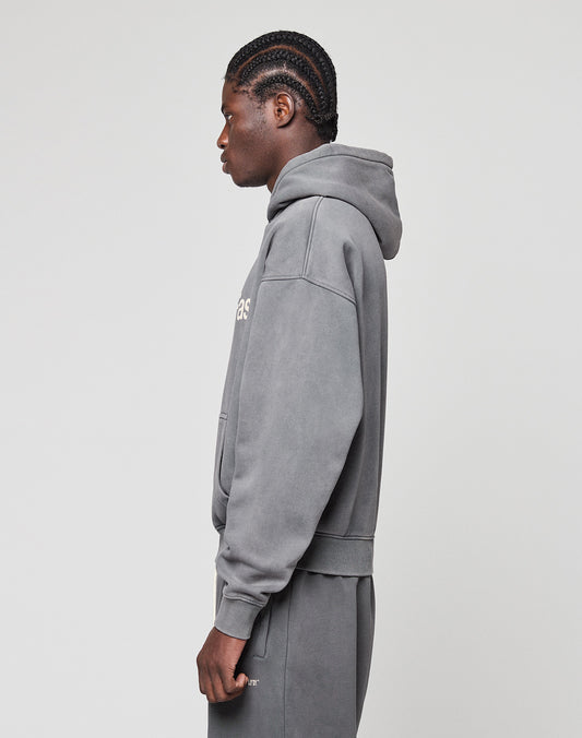 A person with braided hair stands sideways, wearing the LIVE FAST DIE YOUNG Basic +49 Hooded oversized grey sweatshirt with front print and matching cotton sweatpants against a plain light background.