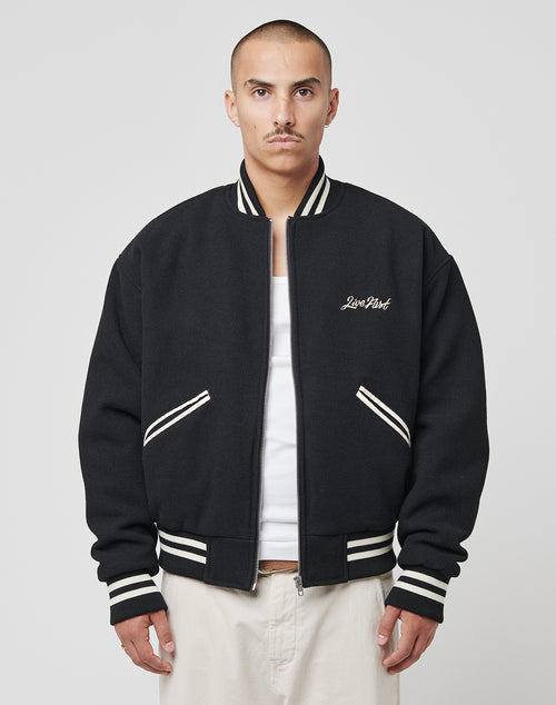 A person with a shaved head wears the LIVE FAST DIE YOUNG Import & Export College Jacket—black polyester with Rückenstick Logo and white contrast stripes on collar, cuffs, and hem—over a white shirt and light pants against a plain background.