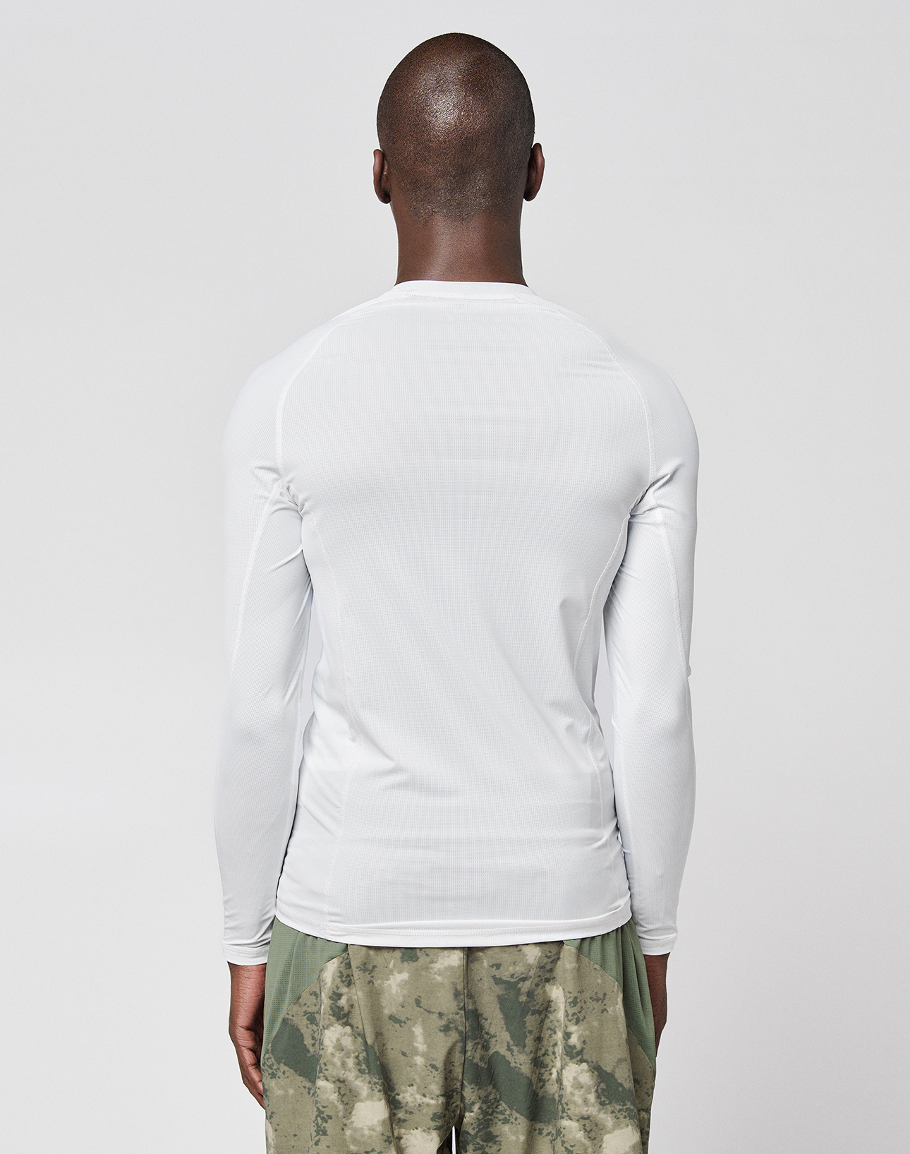 A person with a shaved head stands with their back to the camera, wearing a LIVE FAST DIE YOUNG LFDY Performance Slim Longsleeve in white, featuring mesh insert details, and green camouflage shorts against a plain light background.