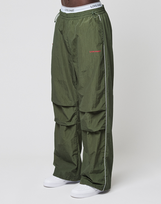 A person wears LFDY Nylon Trackpants by LIVE FAST DIE YOUNG—wide fit, olive green with white piping, LIVE FAST text on waistband and thigh, logo prints—paired with white sneakers, standing against a plain white background.