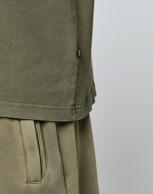 Close-up of a person wearing the olive green LIVE FAST DIE YOUNG Basic 210 Tee, made from 100% Baumwolle. The image shows only the midsection, highlighting the fabric texture and a small black tag on the side seam against a plain background.