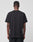 A person with short hair faces away from the camera, wearing a LIVE FAST DIE YOUNG LFDY Blank Tee—regular fit, plain black, short-sleeve, 100% cotton—and black pants against a plain light gray background.