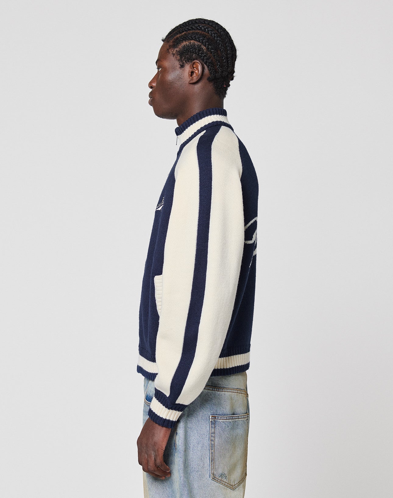 A person with braided hair stands sideways against a gray background, wearing the LIVE FAST DIE YOUNG Boxy Knit Zip Jacket in navy and white with Raglan sleeves, stripes, and an Oversize Fit, paired with light-wash jeans.