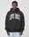 A man wearing the LIVE FAST DIE YOUNG Live Fast College Hooded, an oversize fit black cotton hoodie with bold frontprint, faces the camera against a plain light background.