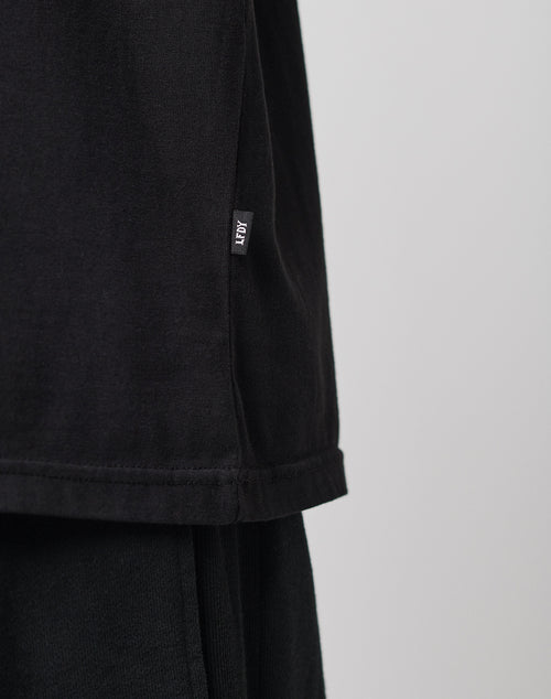 Close-up of the lower side of a black LIVE FAST DIE YOUNG LFDY Blank Tee with a small ADY tag sewn into the side seam, paired with black pants. Neutral, blurred background. Regular fit offers everyday comfort.